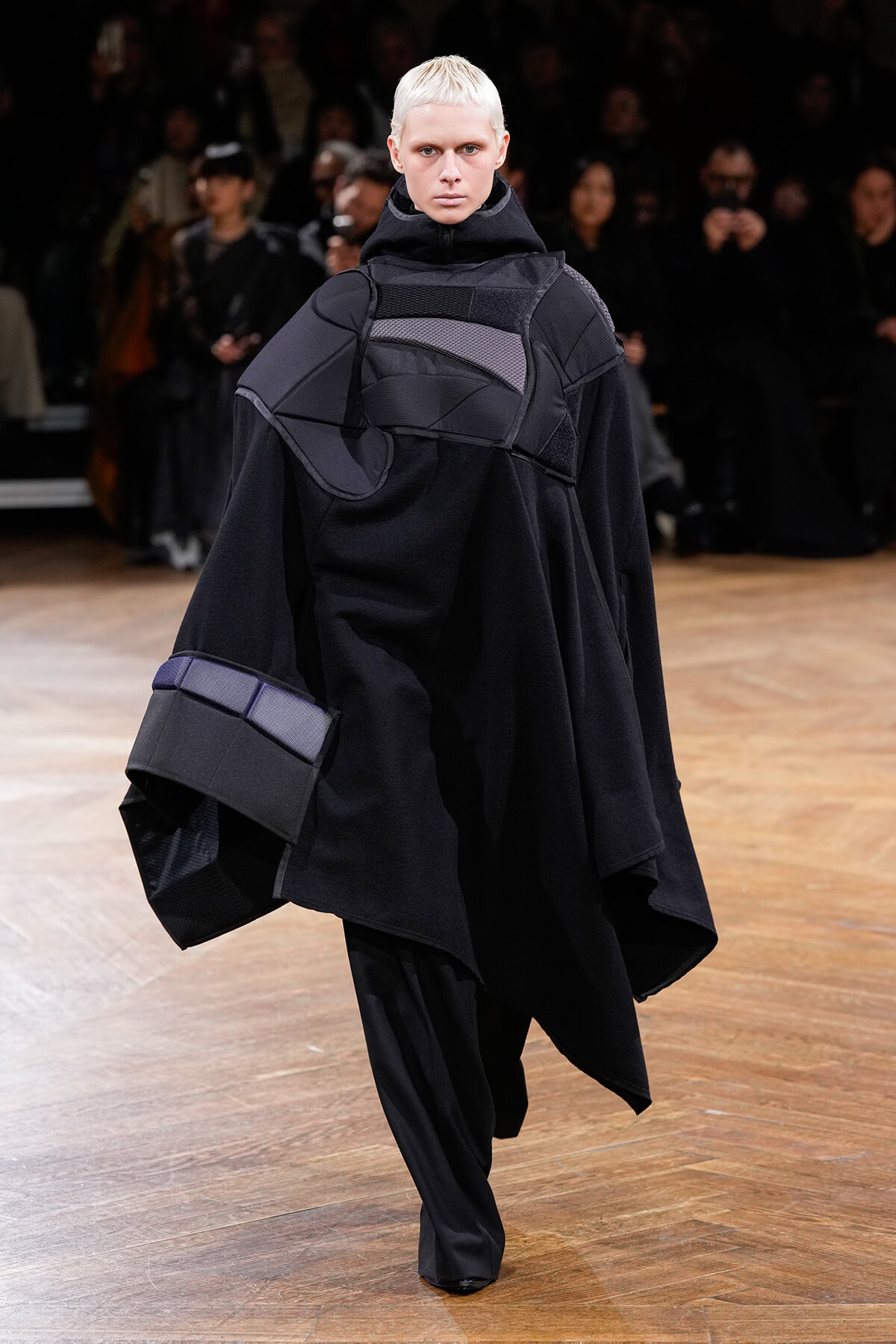 Model walking a runway in an oversized black avant-garde outfit with layered geometric panels, high collar hood, and wide sleeves; dark, minimal styling with a light background audience.