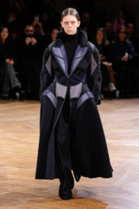 Model wearing a futuristic, padded black and purple geometric coat walking the runway with an audience in the background.