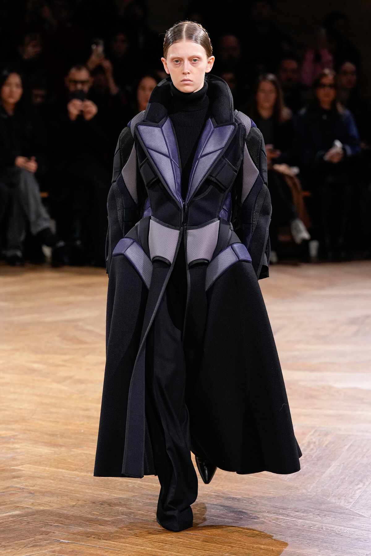 Model wearing a futuristic, padded black and purple geometric coat walking the runway with an audience in the background.