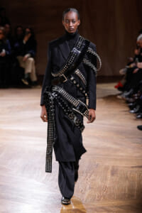 Model walks a dark, tailored suit adorned with multiple metallic studded belts and harness-like straps on a wooden runway.