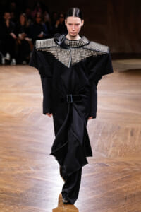 Model on a runway wearing a black oversized coat with large silver studded shoulder panels and a belt.