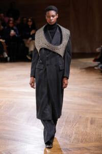 Model walks a runway in a dark pinstripe double-breasted suit with a large metallic bead neckpiece over the shoulders and chest, audience in the background