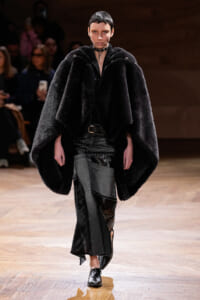 Model walking a runway in a voluminous black fur cape, paired with distressed patchwork pants and black ankle boots; looks directly ahead with a choker necklace as audience watches from the background.
