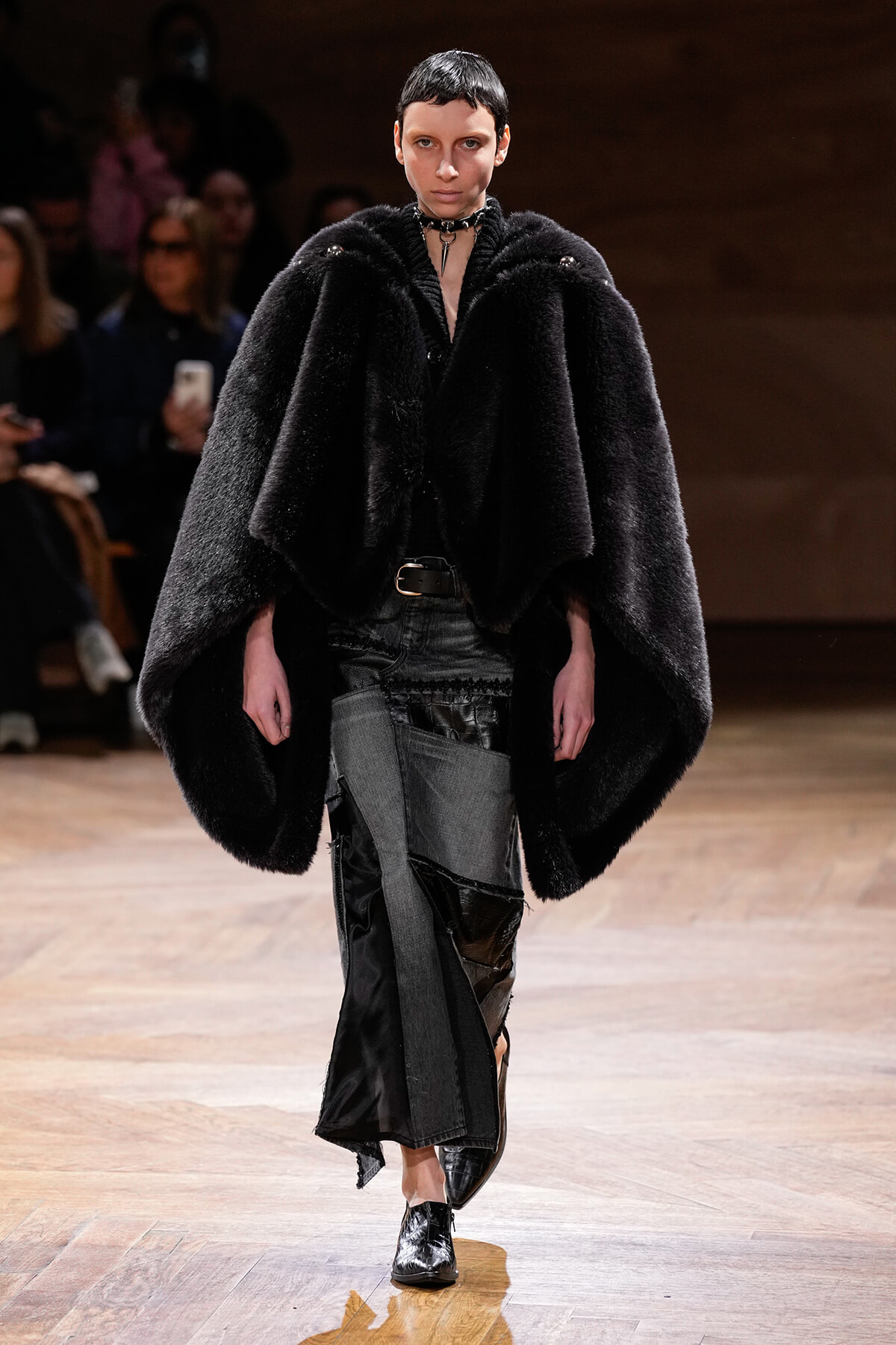 Model walking a runway in a voluminous black fur cape, paired with distressed patchwork pants and black ankle boots; looks directly ahead with a choker necklace as audience watches from the background.