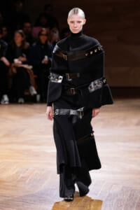 Model on a wooden runway wearing an all-black, layered outfit with wide leather belts and metal studs.