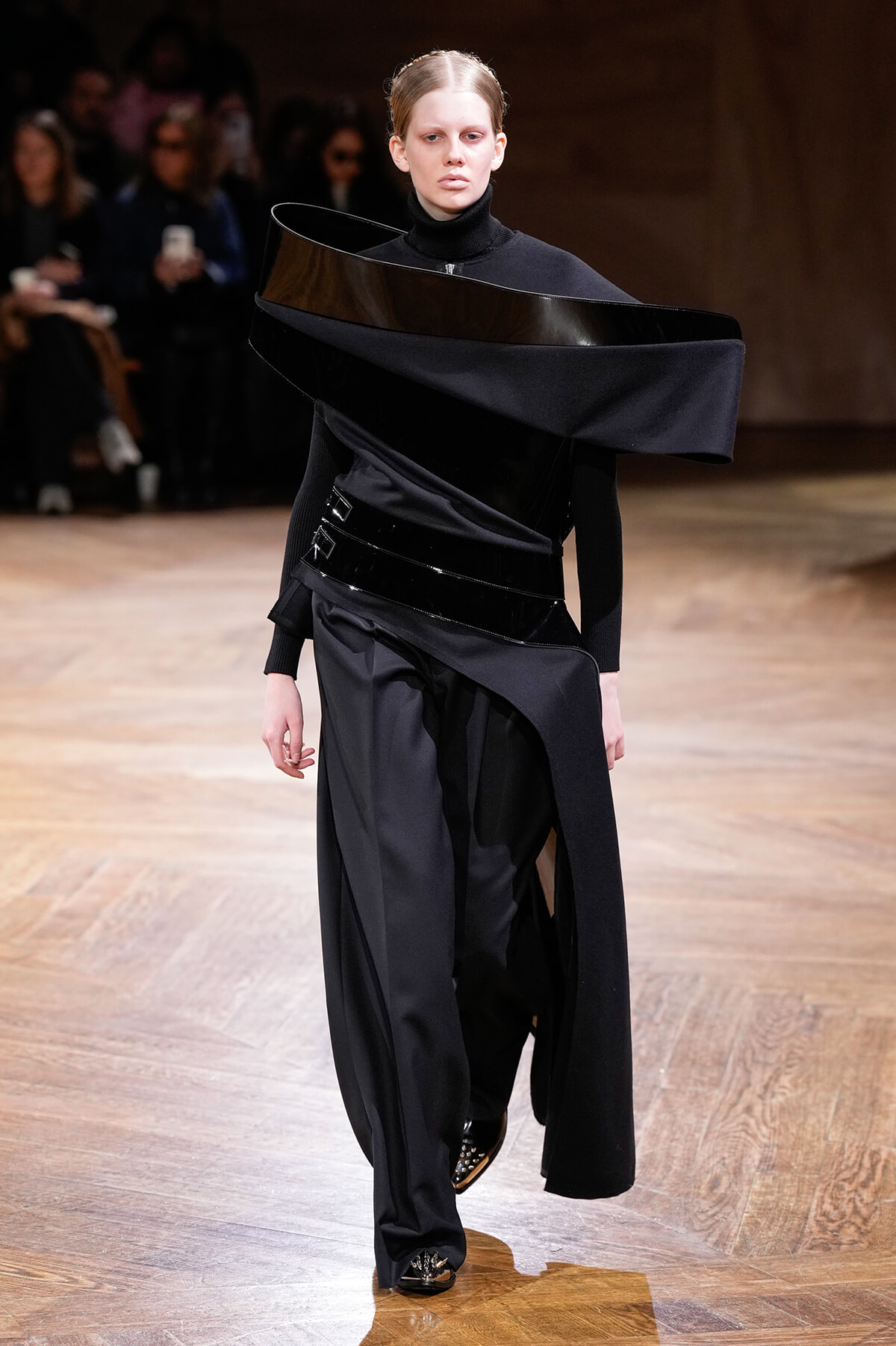 Model on a runway in a black avant-garde outfit with a curved glossy collar wrapping around the shoulders.