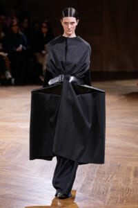 Model walking on a runway in an oversized black coat with a wide belt.
