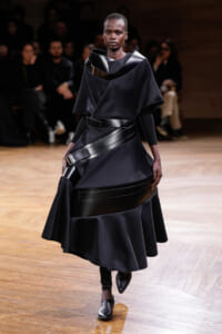 Model on a runway wearing a sculptural black dress with wide leather belts and buckled straps.