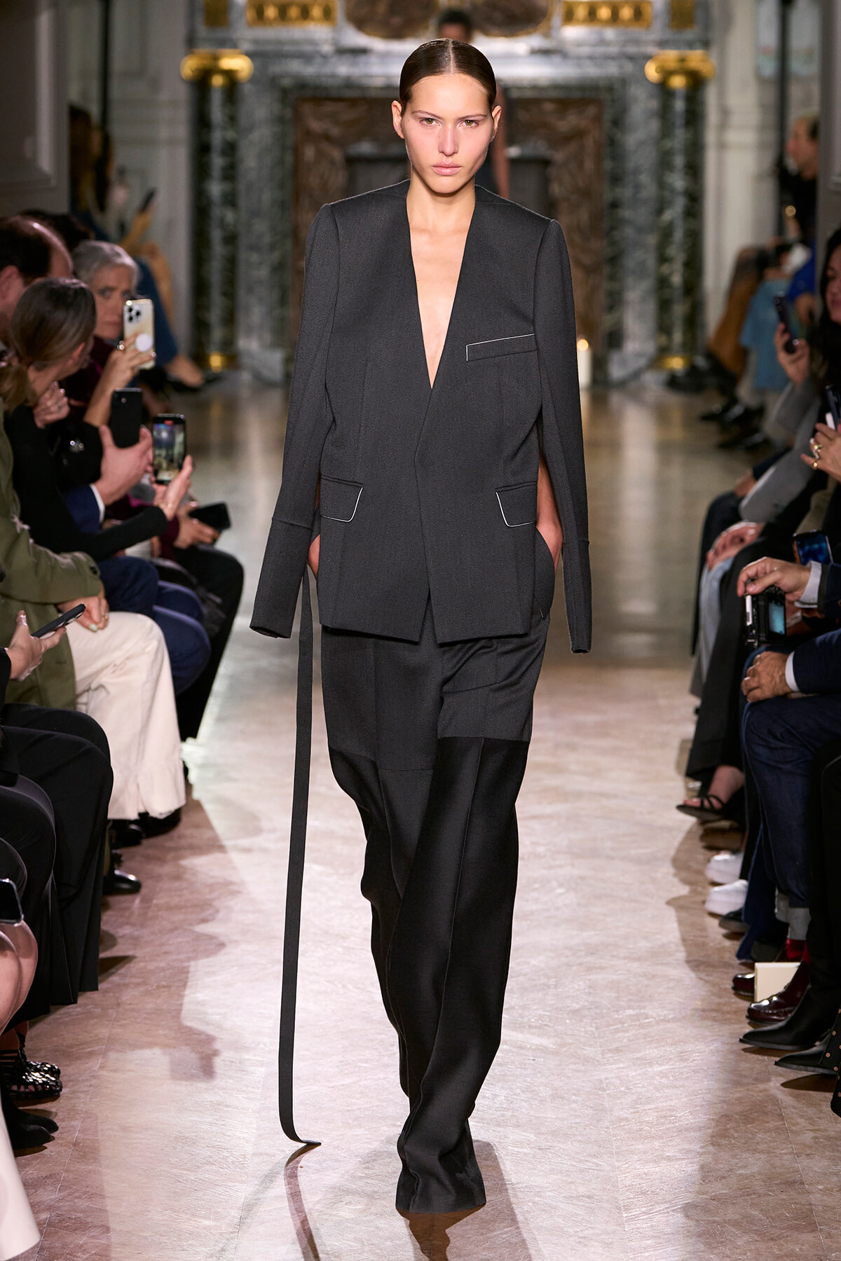 Model walks a dark tailored pantsuit down the runway, audience snapping photos on both sides.