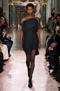 Model walks a fashion runway in a dark gray, asymmetric-shoulder dress with black tights as spectators film and photograph the look.
