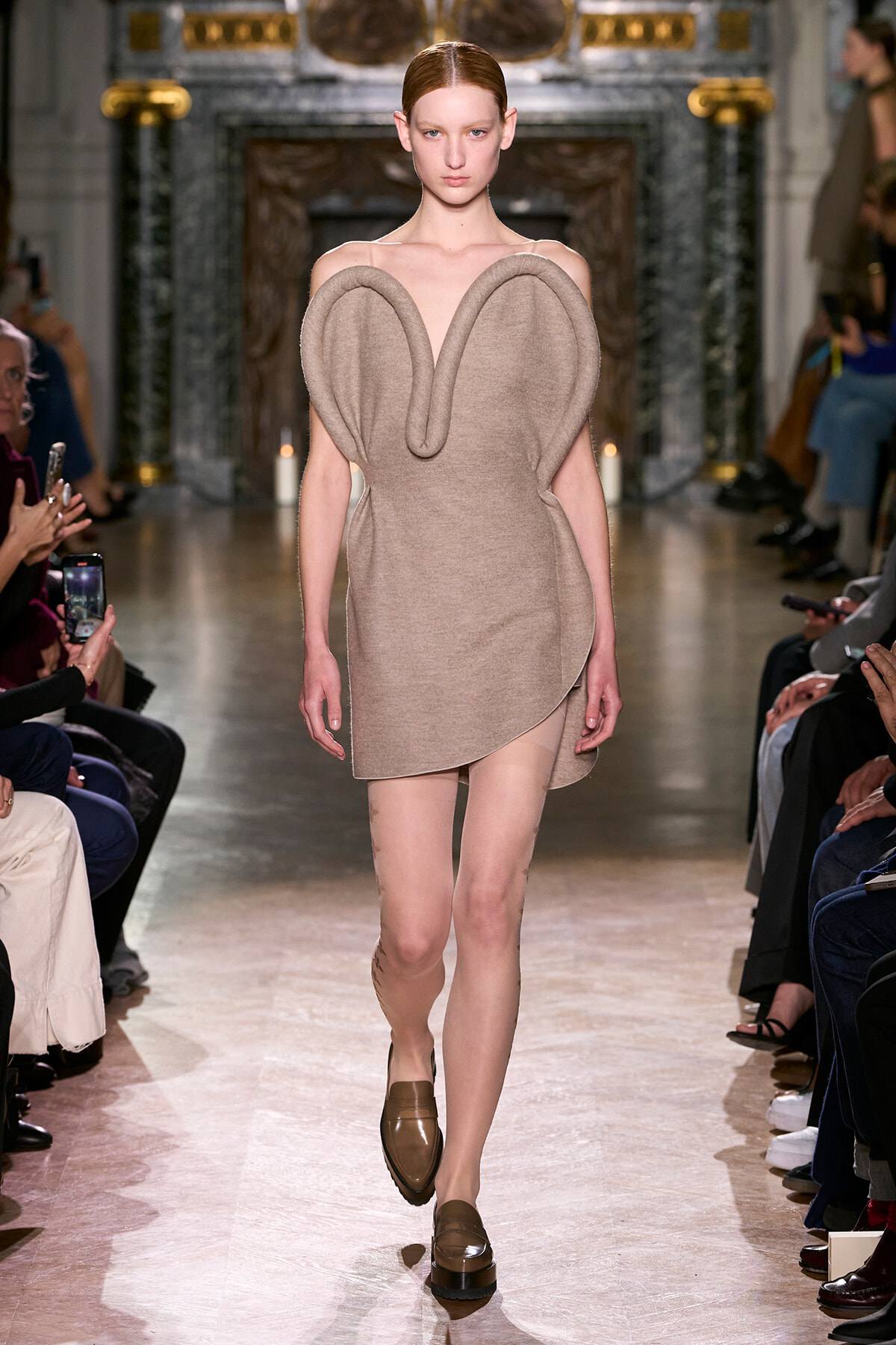 Model walking a runway in a beige sculptural dress with large, rounded shoulder/elbow shapes resembling elephant ears; audience on both sides in a grand hall.