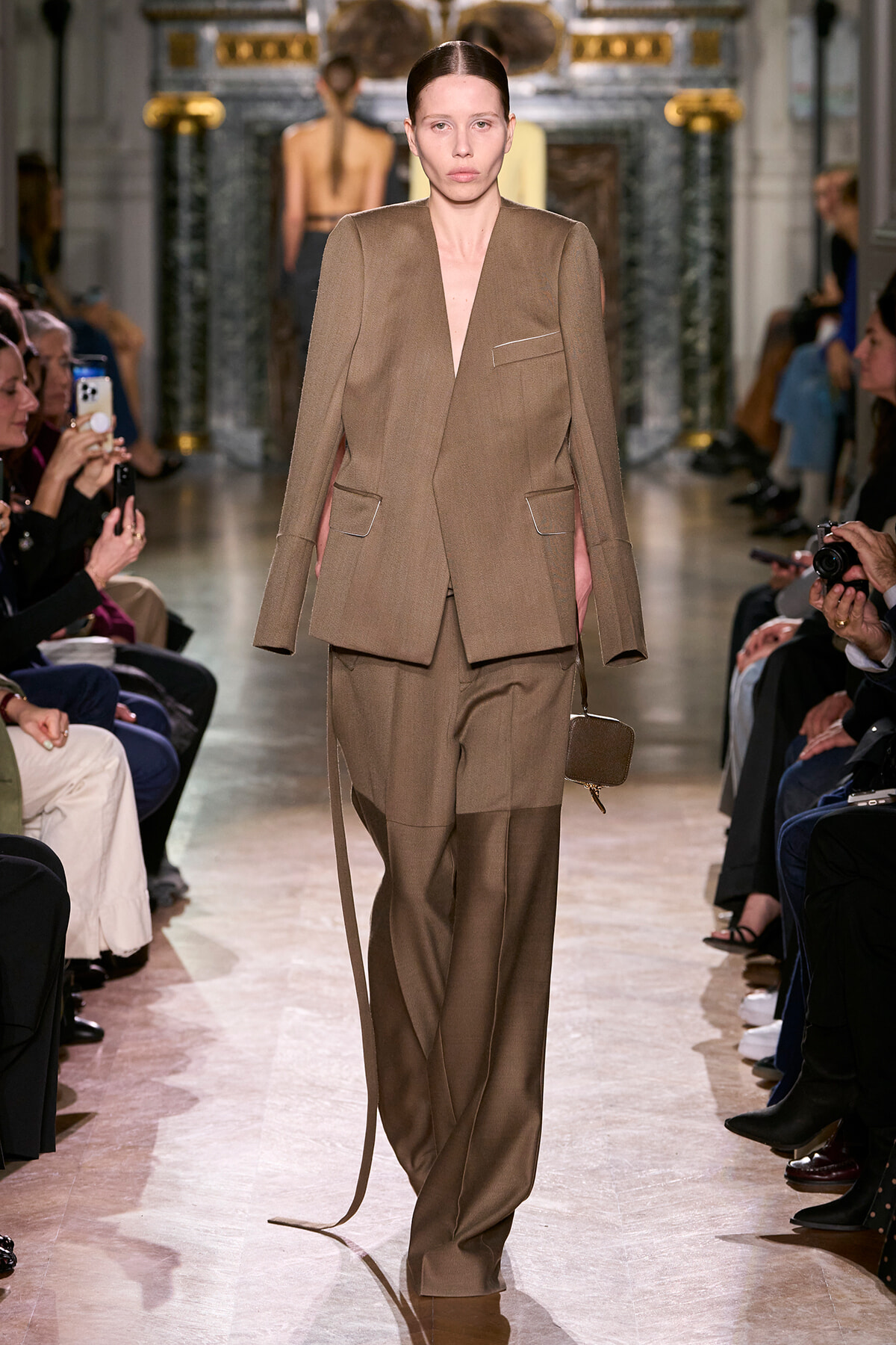 Model on a runway wearing a taupe oversized blazer and matching wide-leg trousers; audience seated on both sides.