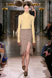 Model walking a fashion show in a pale yellow turtleneck and beige wrap skirt with a front slit, ornate venue in the background.