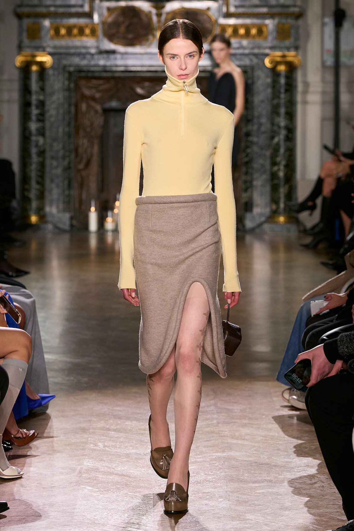 Model walking a fashion show in a pale yellow turtleneck and beige wrap skirt with a front slit, ornate venue in the background.