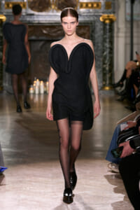 Model on a runway wearing a black sculptural dress with large heart-shaped shoulder panels and black loafers with sheer patterned tights on.