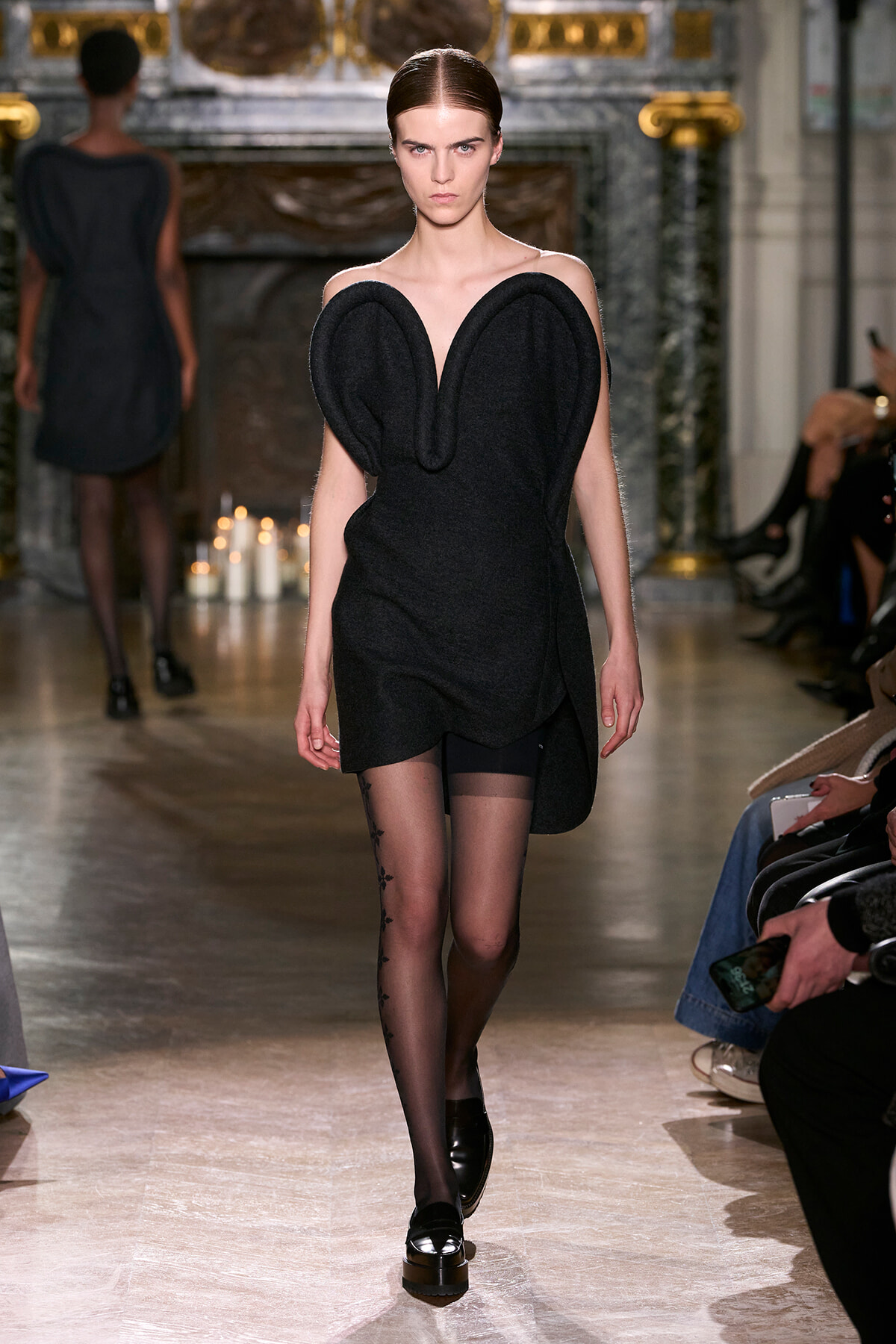 Model on a runway wearing a black sculptural dress with large heart-shaped shoulder panels and black loafers with sheer patterned tights on.