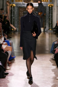 Model walks a runway in a navy turtleneck top and dark split-front skirt, clutch in hand.