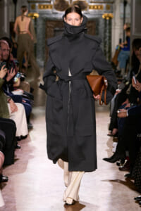 Model walks a fashion runway in an oversized dark charcoal coat with a high collar, tied at the waist, carrying a brown structured handbag; beige trousers peek out underneath.