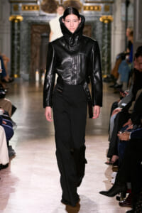 Model walks the runway in a black leather jacket with a high zip collar and black pants.