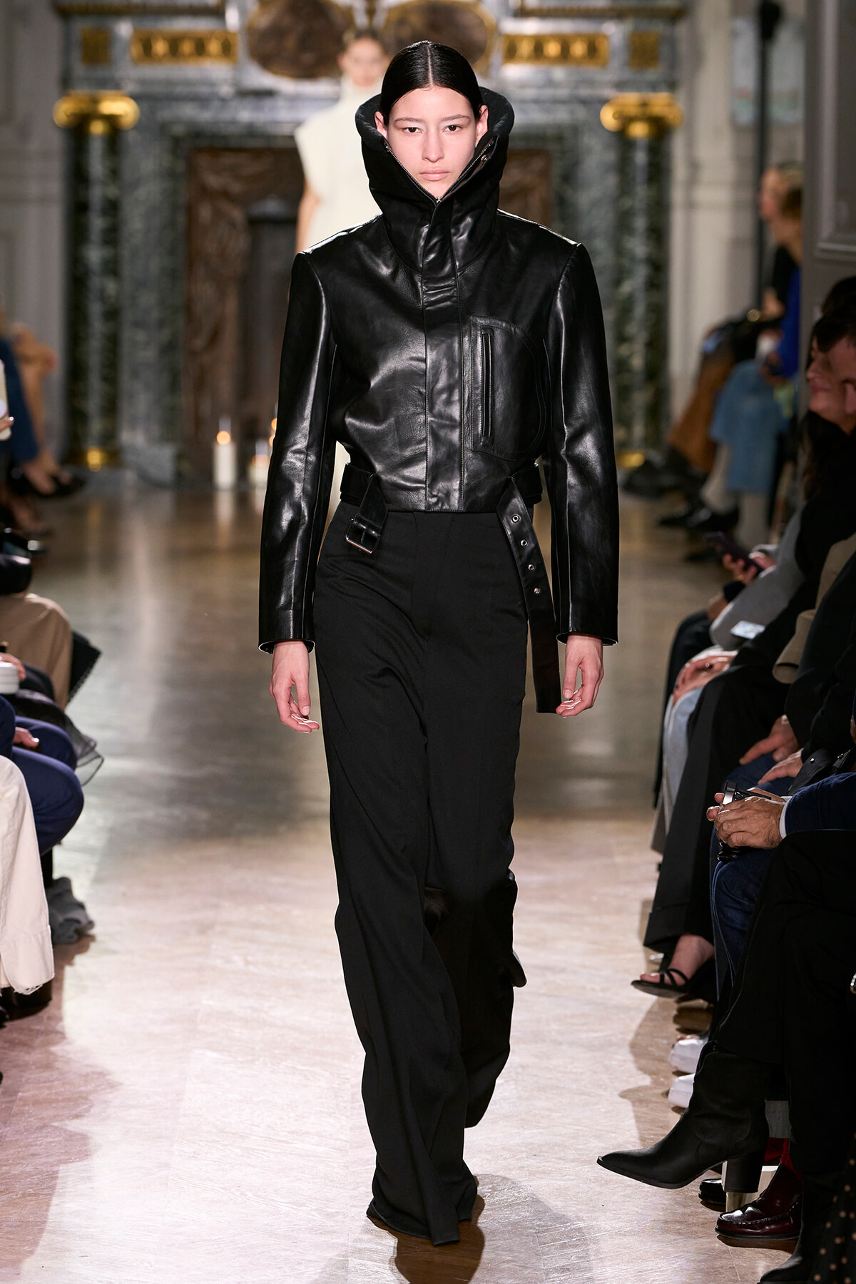 Model walks the runway in a black leather jacket with a high zip collar and black pants.