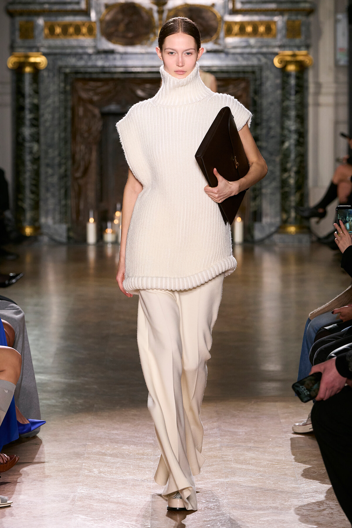 Runway model in an oversized white knit turtleneck sweater with cream wide-leg pants, holding a large black leather portfolio under her arm.