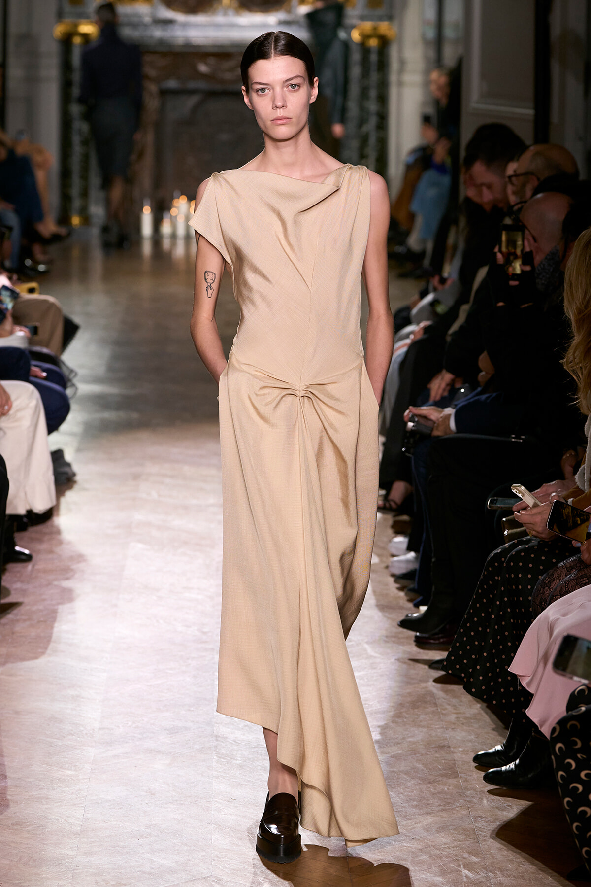 Model walks the runway in a beige draped gown with a gathered waist, flanked by seated audience members.