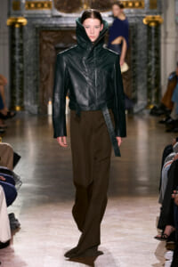 Model walks the runway in a black leather jacket with a tall, oversized collar and brown wide-leg pants.
