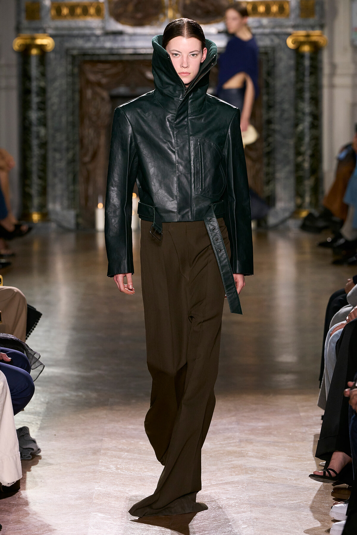 Model walks the runway in a black leather jacket with a tall, oversized collar and brown wide-leg pants.