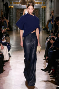 Model walking a fashion runway in a cobalt blue knit poncho and high-waisted navy leather pants, holding a small beige clutch.