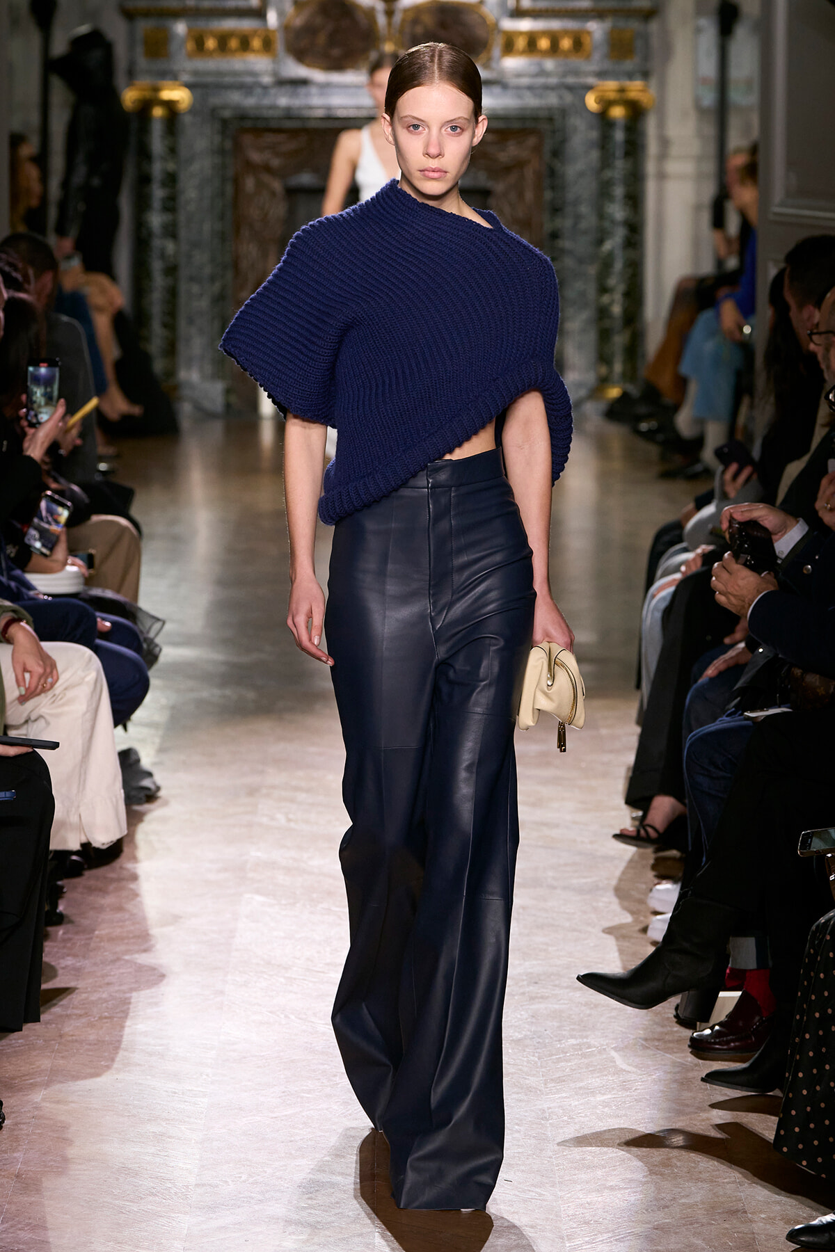 Model walking a fashion runway in a cobalt blue knit poncho and high-waisted navy leather pants, holding a small beige clutch.