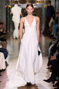 Model walks the runway in a floor-length white sleeveless gown with a deep V neckline and side pockets, audience on both sides.