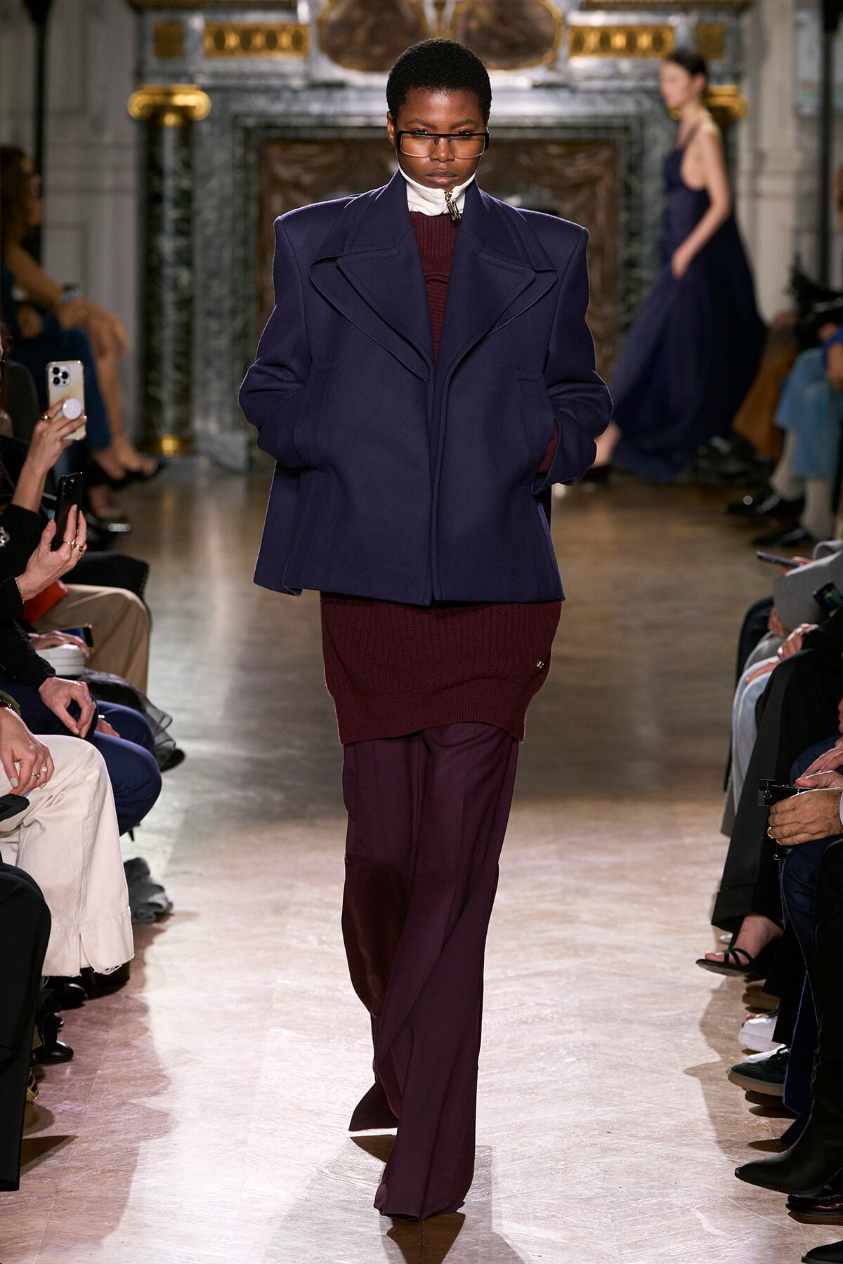 Model walks the runway in an oversized navy blazer layered over a burgundy knit dress and matching pants, flanked by seated audience in a grand venue.