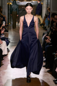 Model wearing a navy satin gown with a plunging V-neck walks the runway as photographers and spectators line the catwalk.