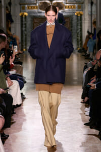 Model on a runway wearing an oversized navy coat over a camel knit top and beige wide-leg pants, audience on both sides watching the show.