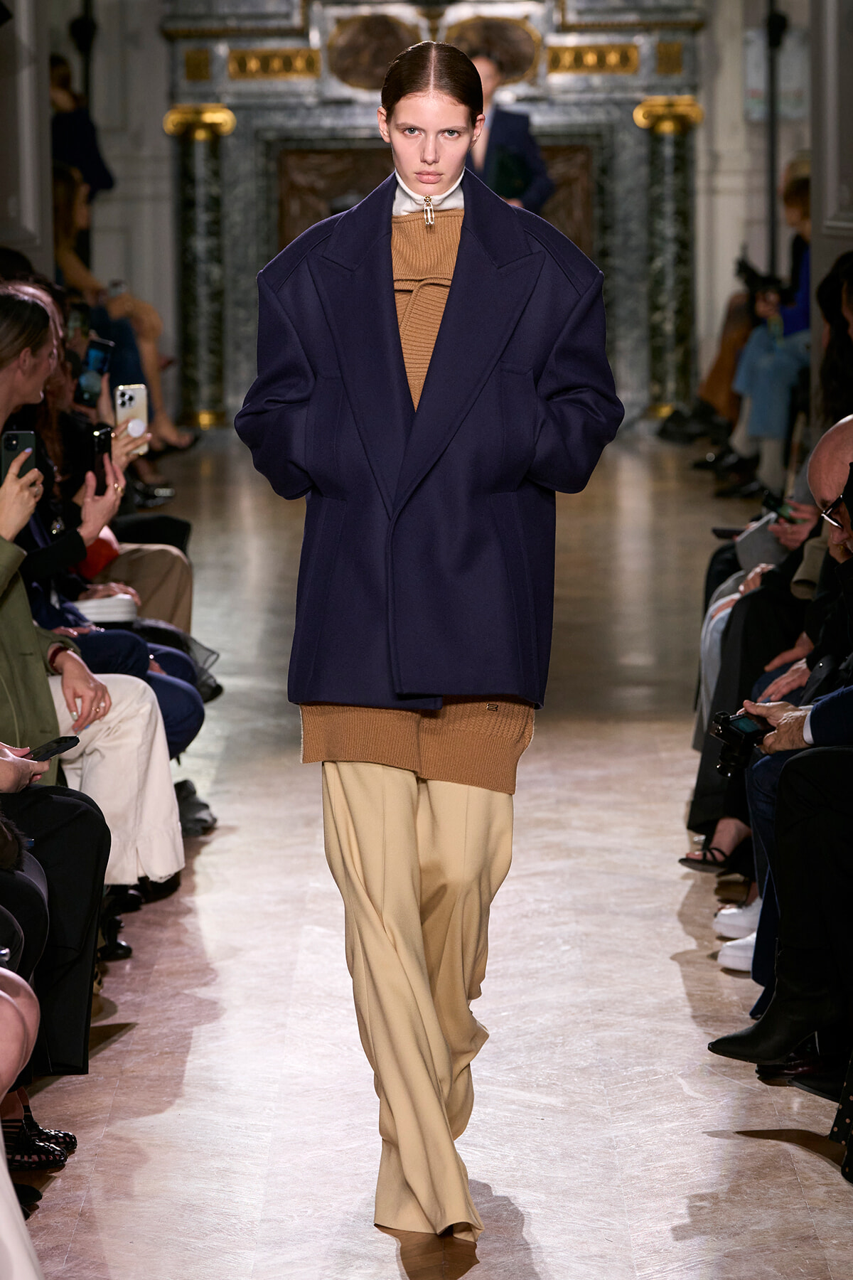 Model on a runway wearing an oversized navy coat over a camel knit top and beige wide-leg pants, audience on both sides watching the show.