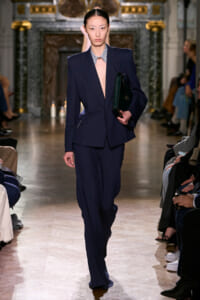 Model walks the runway in a navy tailored suit with an open blazer and striped shirt, clutch in hand, audience in the background.