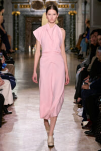 Runway model in a pale pink draped dress walking the catwalk with spectators seated on both sides.
