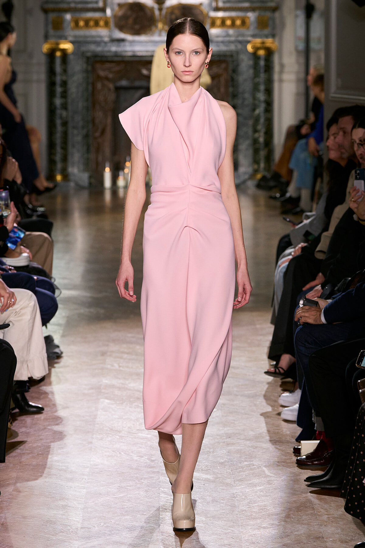 Runway model in a pale pink draped dress walking the catwalk with spectators seated on both sides.