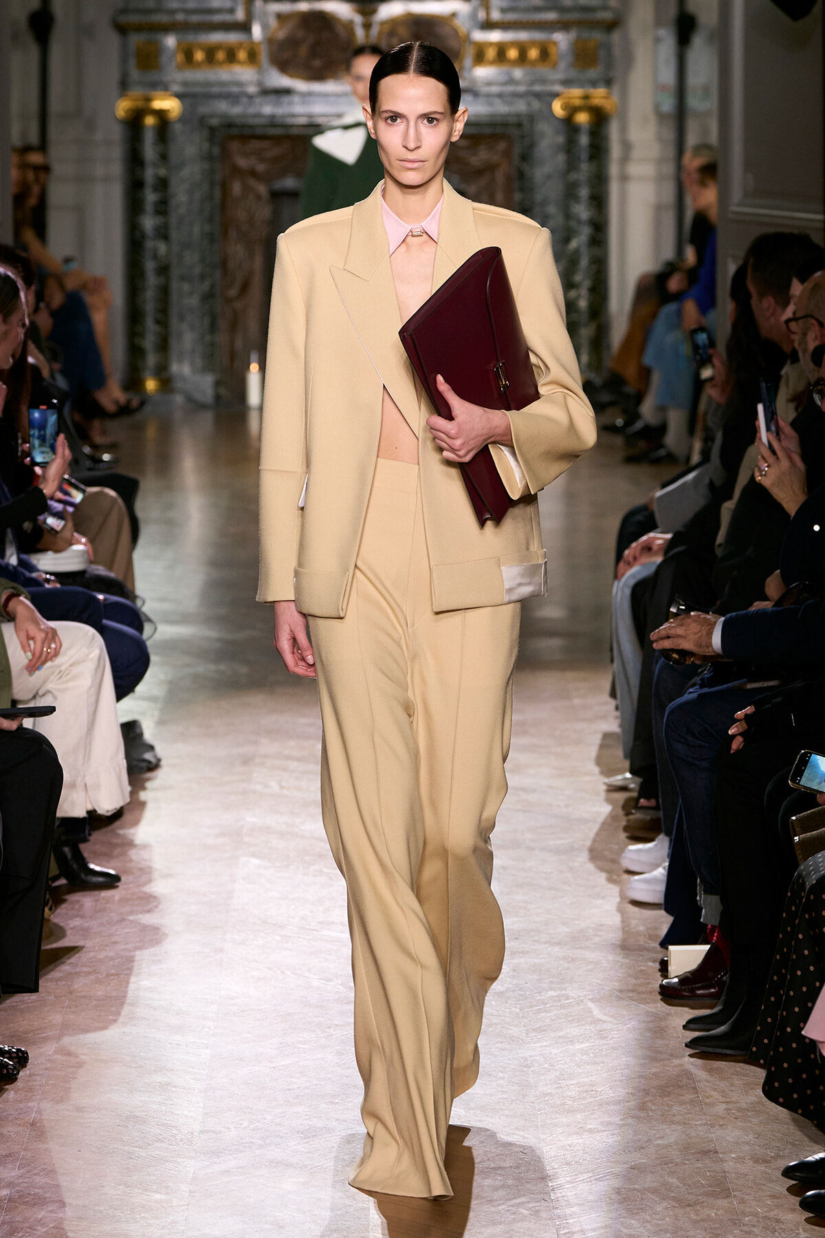 Model walks a fashion-show runway in a beige tailored suit with pale pink shirt, carrying a burgundy clutch. Audience along the sides watch.