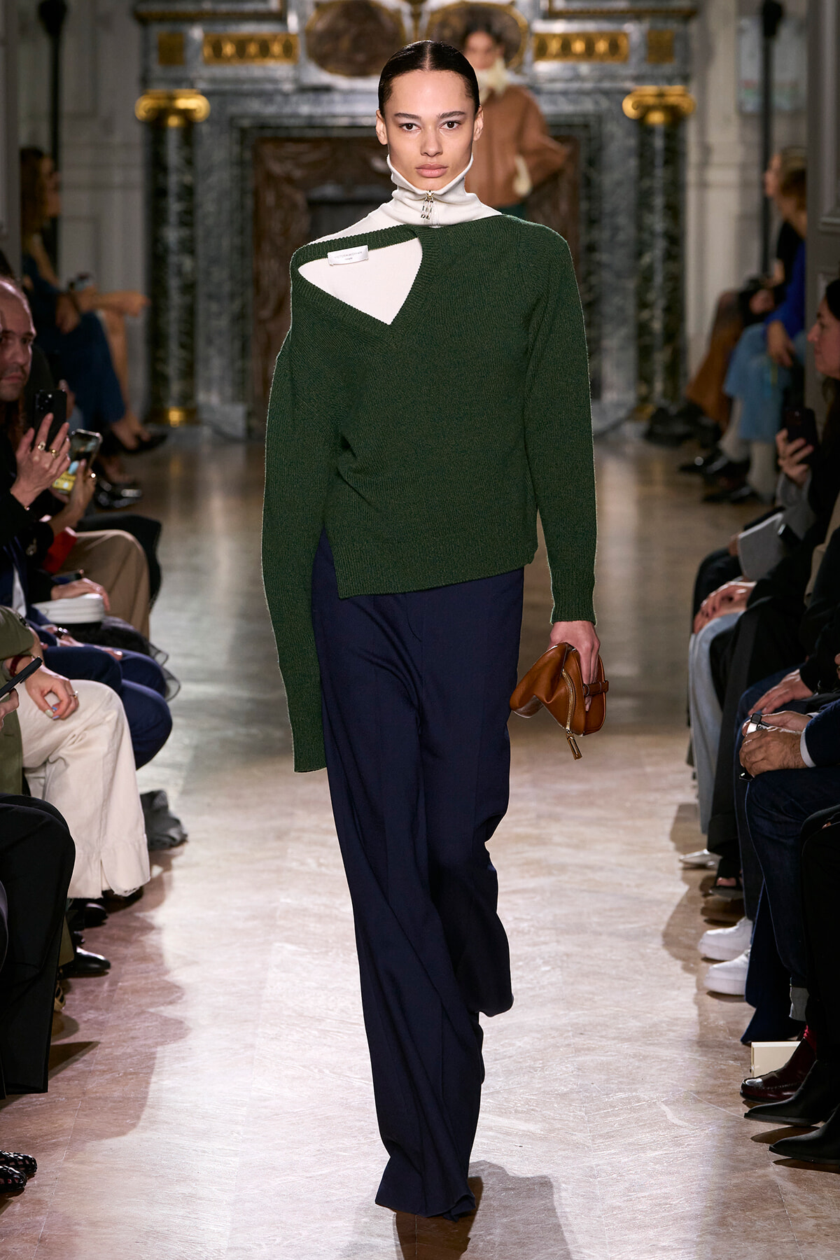 Model walks the runway in a dark green knit sweater over a white turtleneck and navy trousers, holding a brown clutch.