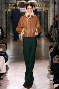 Model on a runway wearing a tan shearling jacket with cream fleece trim and green wide-leg pants, audience visible on both sides.
