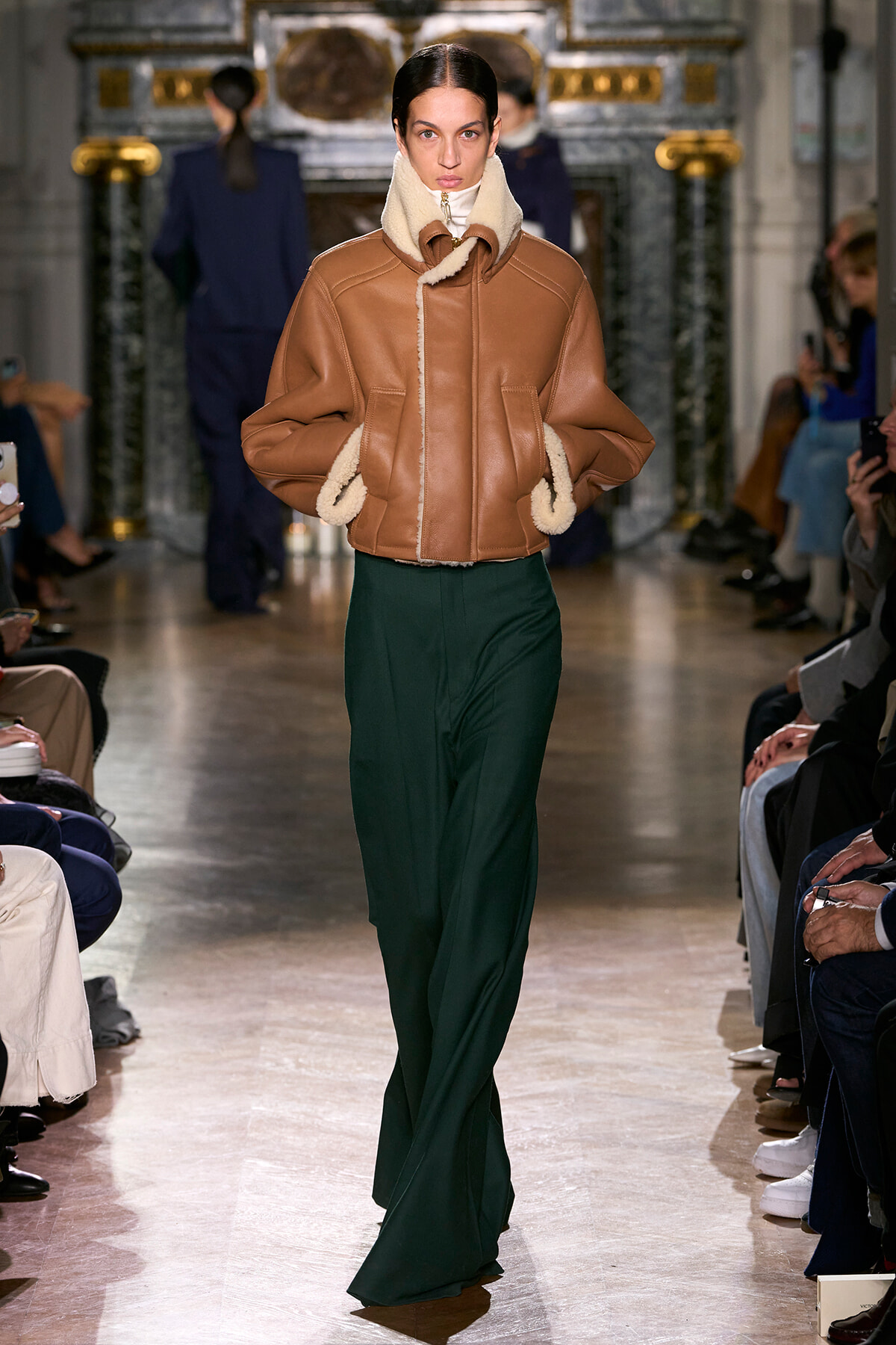 Model on a runway wearing a tan shearling jacket with cream fleece trim and green wide-leg pants, audience visible on both sides.