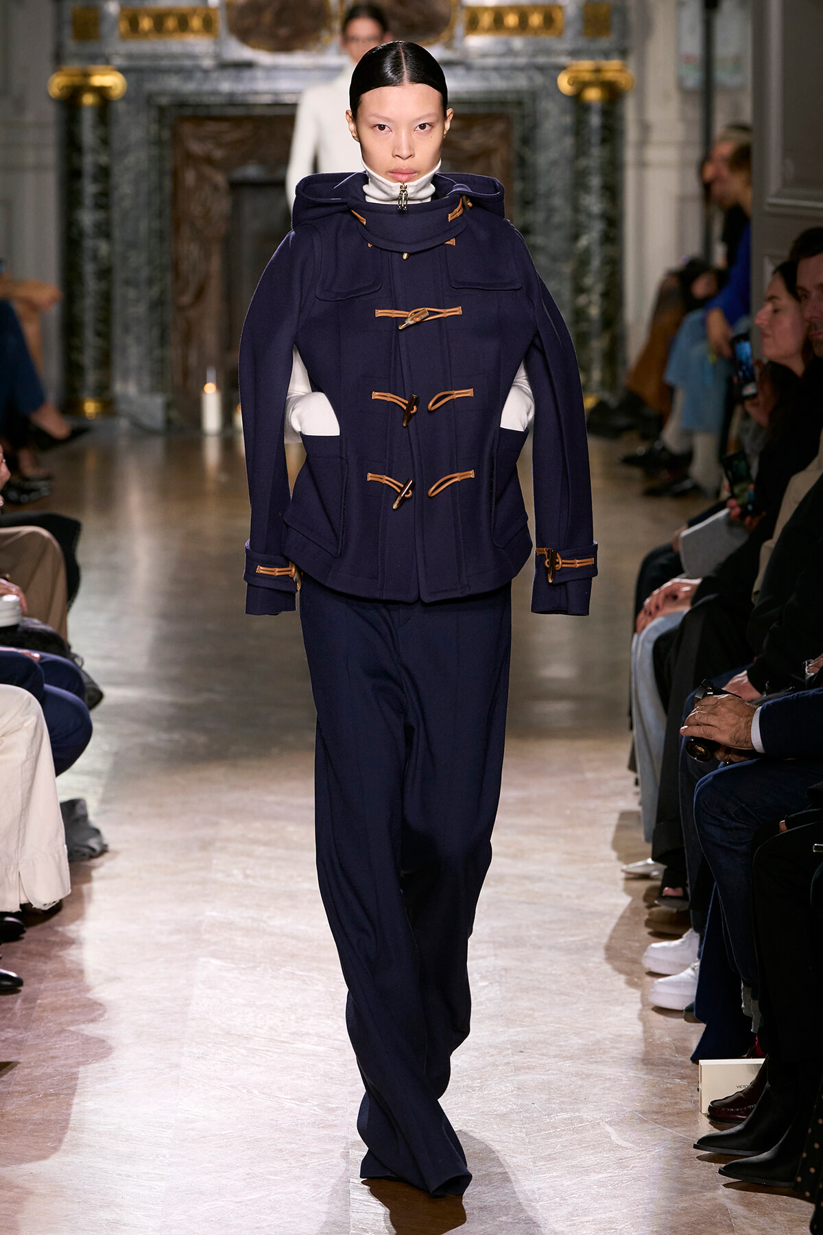 Model walks a fashion runway wearing a navy duffle coat with tan toggles and hooded cape detail.