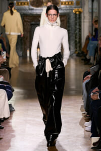 Model walks the runway in a white turtleneck top and glossy black pants with a beige belt, audience seated on both sides.
