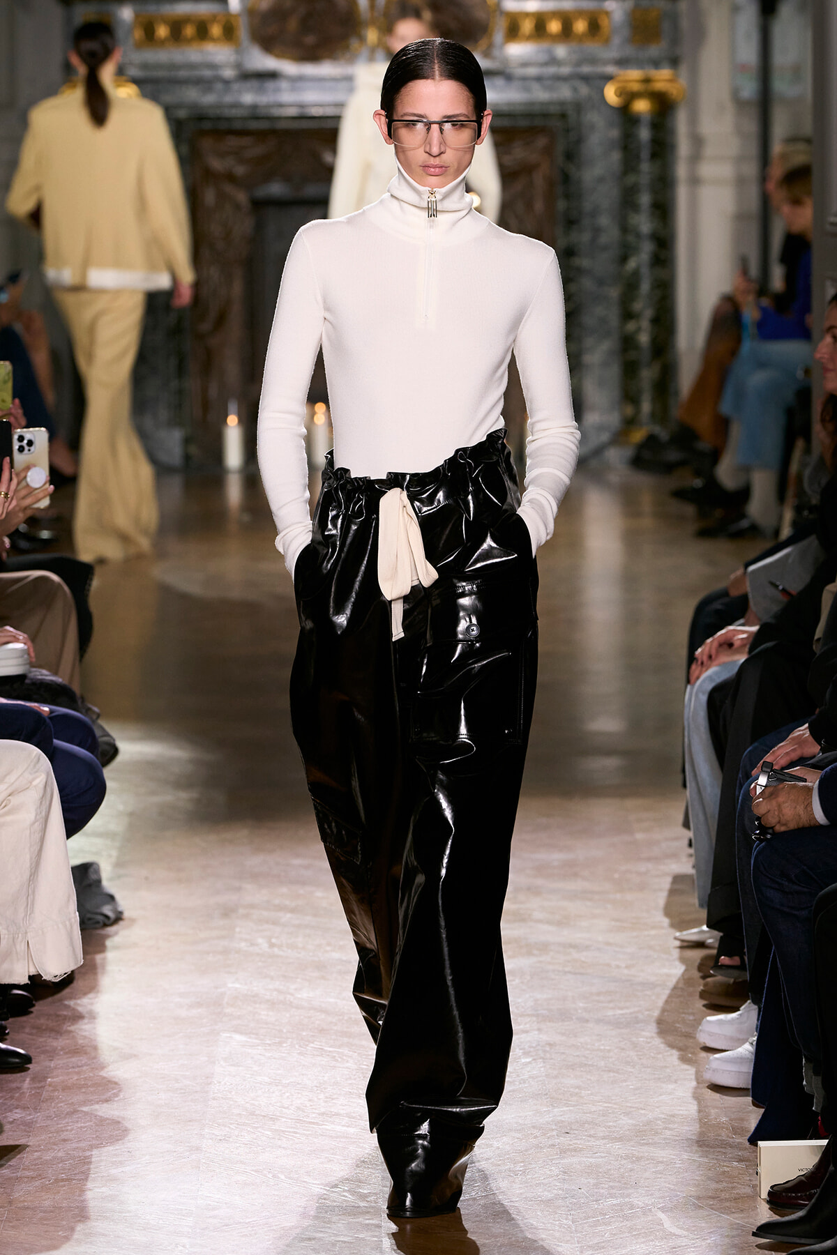 Model walks the runway in a white turtleneck top and glossy black pants with a beige belt, audience seated on both sides.