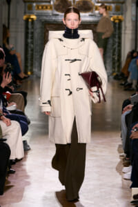 Model walks the runway in a cream oversized coat with toggle closures, holding a burgundy bag.