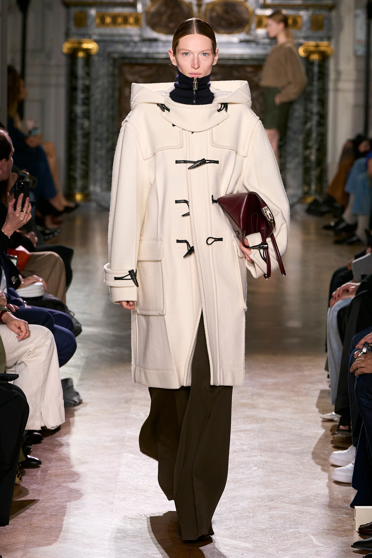 Model walks the runway in a cream oversized coat with toggle closures, holding a burgundy bag.