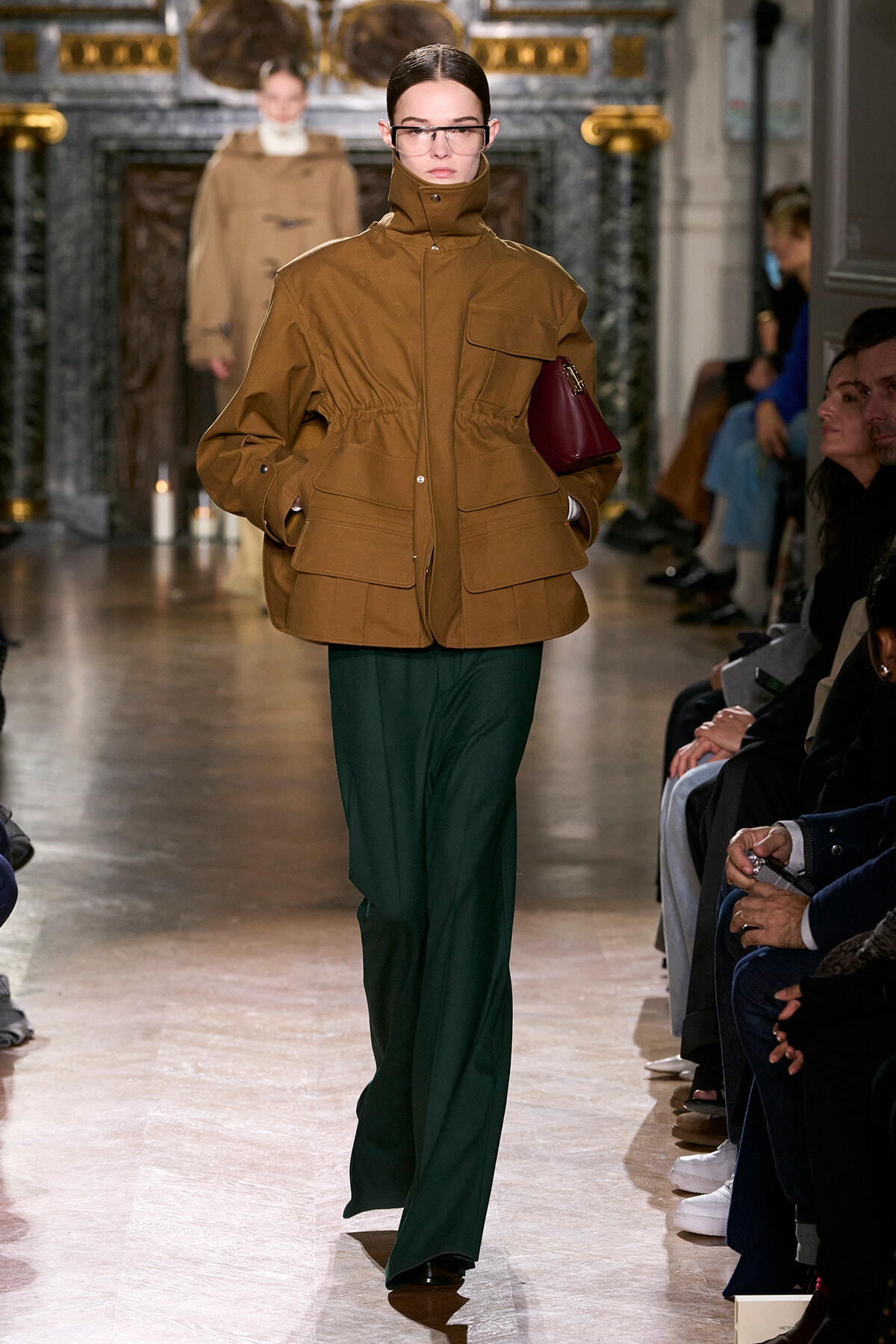 Model walking the runway in a brown padded high-collar jacket with a burgundy handbag, paired with dark green trousers, as audience watches.