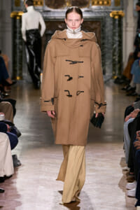 Model walks a runway in a camel duffle coat with large collar and black toggles, holding a small clutch.