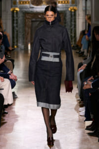Model walks a runway in a dark denim dress with a high collar and a wide belt featuring a gray pouch at the waist.